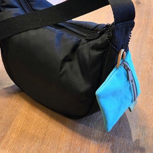 May include: A black nylon shoulder bag with a curved silhouette, a black zip closure, and a wide black strap. A small, rectangular, bright turquoise fabric pouch with a grey zip is attached to the side with a black beaded chain.