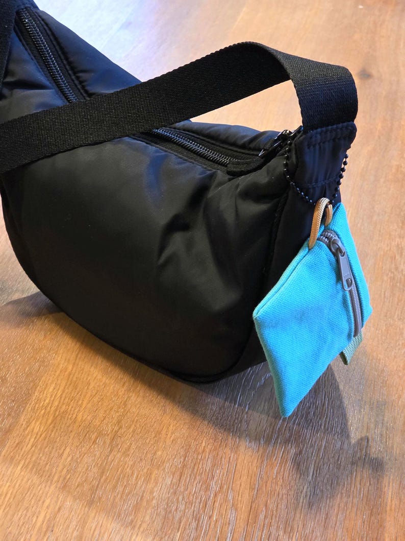 May include: A black nylon shoulder bag with a curved silhouette, a black zip closure, and a wide black strap. A small, rectangular, bright turquoise fabric pouch with a grey zip is attached to the side with a black beaded chain.