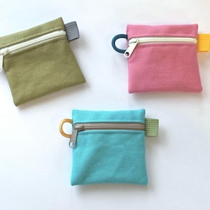 May include: Three square canvas zip pouches in olive green, pink, and turquoise. Each pouch features a white or grey zip, a small circular pull tab on the left side, and a small rectangular fabric tab on the right side in contrasting colours.