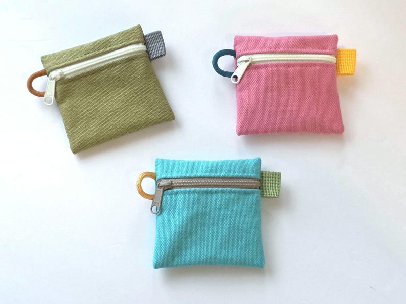 May include: Three square canvas zip pouches in olive green, pink, and turquoise. Each pouch features a white or grey zip, a small circular pull tab on the left side, and a small rectangular fabric tab on the right side in contrasting colours.