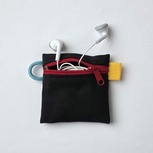 May include: A small black square pouch with a red zip closure, a light blue cord loop on the left side, and a yellow fabric tab on the right side. White wired earphones are tucked inside the open zip compartment of the pouch.