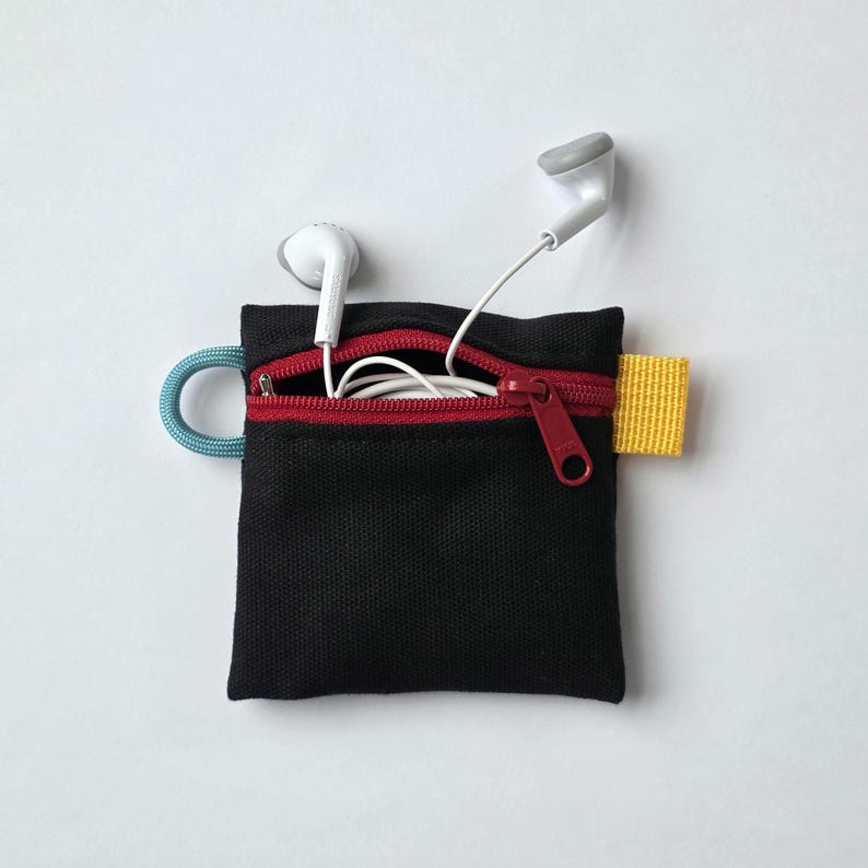 May include: A small black square pouch with a red zip closure, a light blue cord loop on the left side, and a yellow fabric tab on the right side. White wired earphones are tucked inside the open zip compartment of the pouch.
