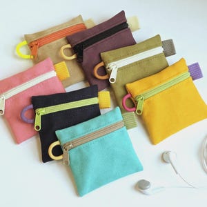 May include: A collection of seven small, square canvas zipper pouches in various colors including tan, dark purple, olive green, pink, black, yellow, and turquoise. Each pouch features a contrasting zipper, a side loop, and a small fabric tab.