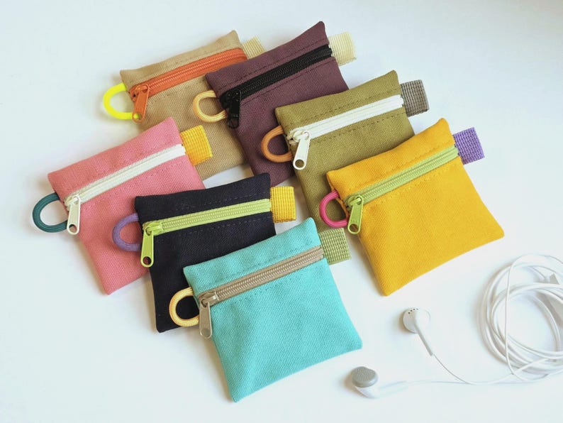 May include: A collection of seven small, square canvas zip pouches in various colours including tan, dark purple, olive green, pink, black, yellow, and turquoise. Each pouch features a contrasting zip, a side loop, and a small fabric tab.