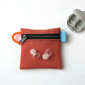 May include: A square, rust-coloured canvas pouch featuring a black zip closure, an orange side loop, and a light blue side tab. Two clear, circular silicone earplugs are positioned on the front of the pouch, with a grey plastic case nearby.