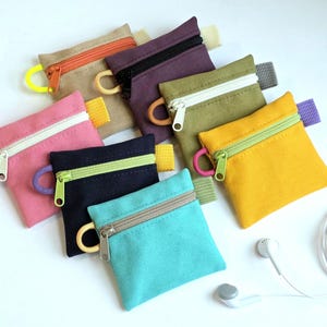 May include: A collection of small, colorful zippered pouches in various shades, including pink, teal, yellow, and navy. Each pouch has a contrasting zipper and a small loop. Earbuds are visible in the lower right corner.