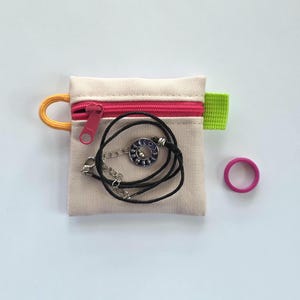May include: A small beige canvas zipped pouch with a bright pink zip, an orange cord loop, and a lime green side tab. Inside the pouch is a black cord necklace with a circular silver pendant featuring blue moon phases. A solid pink ring.