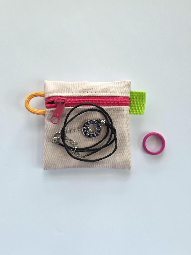 May include: A small beige canvas zipped pouch with a bright pink zip, an orange cord loop, and a lime green side tab. Inside the pouch is a black cord necklace with a circular silver pendant featuring blue moon phases. A solid pink ring.
