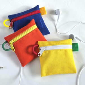 May include: A collection of colorful, small zippered pouches in blue, orange, and yellow. Each pouch has a contrasting zipper and a loop for attaching to a keychain. White earbuds and a headphone jack are also present, suggesting the pouches are for storing headphones.