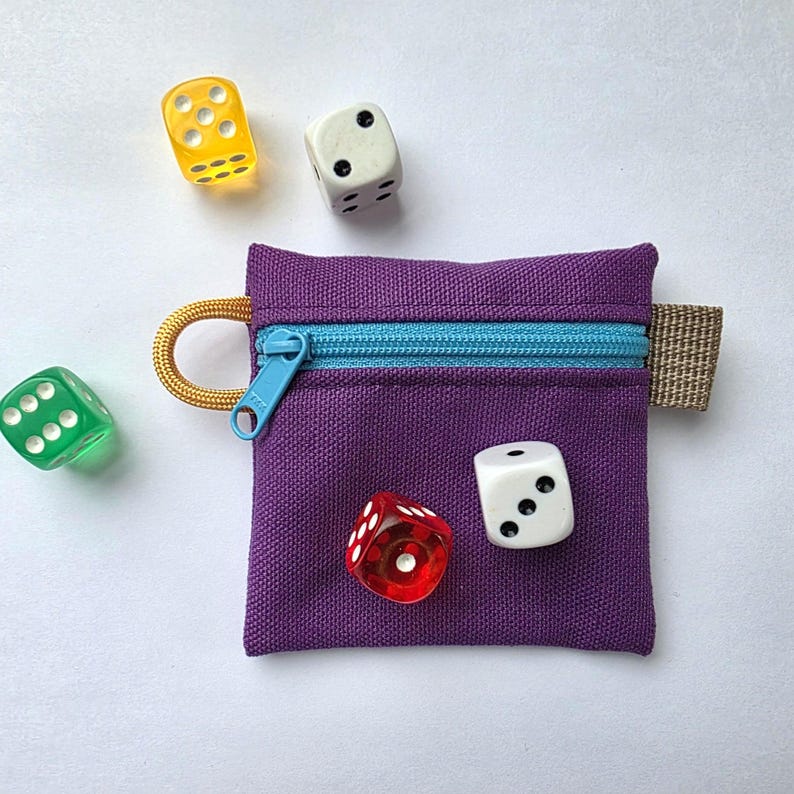 May include: A square purple fabric pouch featuring a bright blue zip, a gold cord loop on the left side, and a beige fabric tab on the right. Four six-sided dice in yellow, white, green, and red are positioned around the pouch.