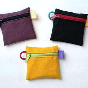 May include: Three square zipped pouches in purple, black, and yellow. Each pouch features a top zip closure, a small side loop, and a contrasting fabric tab on the opposite side. The zips and loops are in various colours including red, green, and orange.