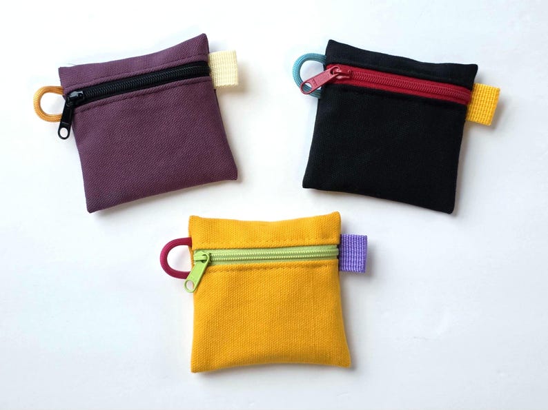 May include: Three square zipped pouches in purple, black, and yellow. Each pouch features a top zip closure, a small side loop, and a contrasting fabric tab on the opposite side. The zips and loops are in various colours including red, green, and orange.