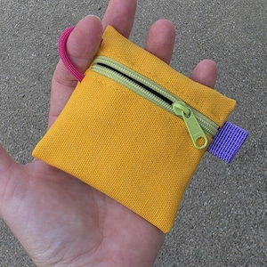 May include: A small, square, yellow canvas pouch featuring a lime green zippered closure with a matching lime green zipper pull. The pouch includes a bright pink cord loop attached to the top left corner and a purple fabric tab on the right side.