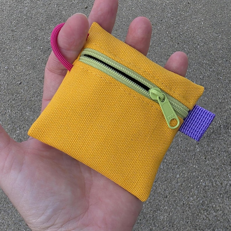 May include: A small, square, yellow canvas pouch featuring a lime green zippered closure with a matching lime green zipper pull. The pouch includes a bright pink cord loop attached to the top left corner and a purple fabric tab on the right side.