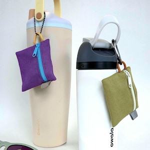 May include: A beige Owala water bottle with a yellow handle and a purple square zipped pouch attached, and a white Owala water bottle with a grey lid and an olive green square zipped pouch attached, with light green sunglasses in the foreground.