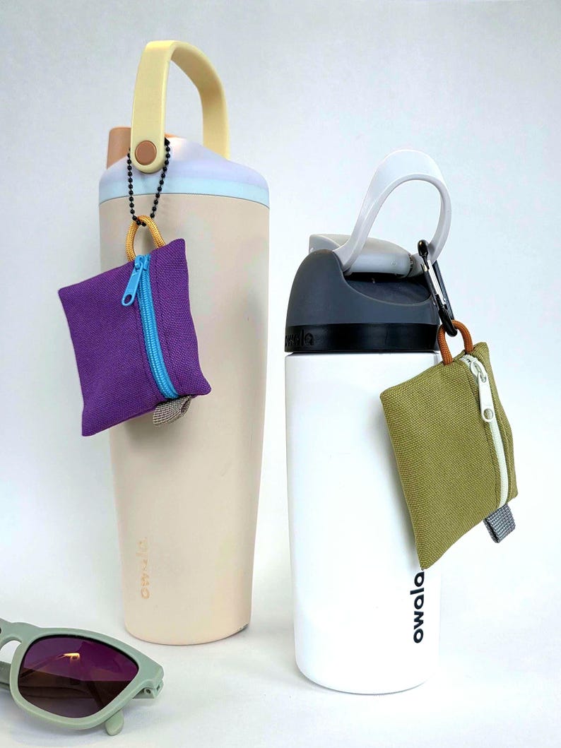 May include: A beige Owala water bottle with a yellow handle and a purple square zipped pouch attached, and a white Owala water bottle with a grey lid and an olive green square zipped pouch attached, with light green sunglasses in the foreground.