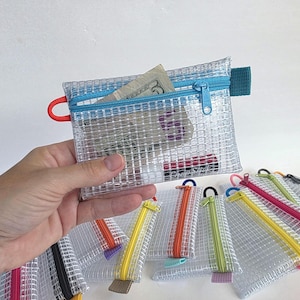 May include: A rectangular, clear, reinforced mesh zipper pouch with a bright blue zipper closure. The pouch features a small red loop on the left side and a teal fabric tab on the right. Inside, a five-dollar bill and a small tube are visible.