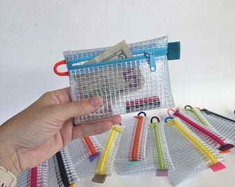 May include: A rectangular, clear, reinforced mesh zip pouch with a bright blue zip closure. The pouch features a small red loop on the left side and a teal fabric tab on the right. Inside, a five-pound note and a small tube are clearly visible.