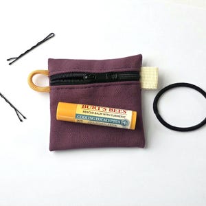 May include: A small, square, plum-coloured canvas zipped pouch with a yellow cord loop and a white fabric tab. A yellow Burt's Bees Cooling Eucalyptus lip balm tube rests on the pouch, accompanied by two black bobby pins and a black hair tie.