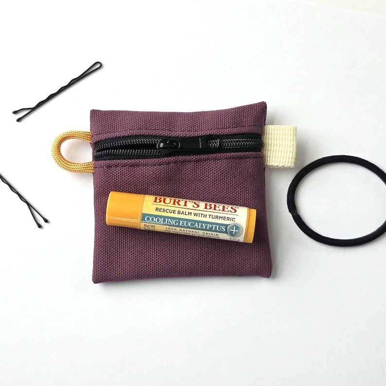 May include: A small, square, plum-coloured canvas zipped pouch with a yellow cord loop and a white fabric tab. A yellow Burt's Bees Cooling Eucalyptus lip balm tube rests on the pouch, accompanied by two black bobby pins and a black hair tie.