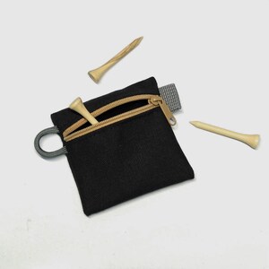 May include: A small black square zipped pouch with a tan zip tape and a grey side loop. Two light-coloured wooden golf tees are positioned near the pouch, and one additional wooden golf tee is partially inserted into the open zip compartment.