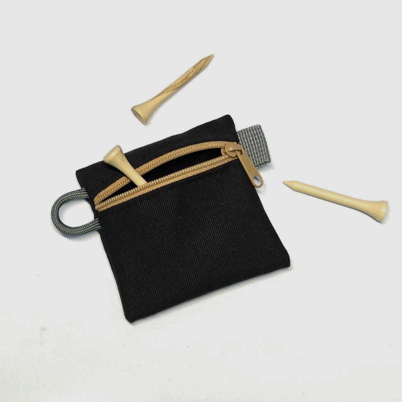 May include: A small black square zipped pouch with a tan zip tape and a grey side loop. Two light-coloured wooden golf tees are positioned near the pouch, and one additional wooden golf tee is partially inserted into the open zip compartment.