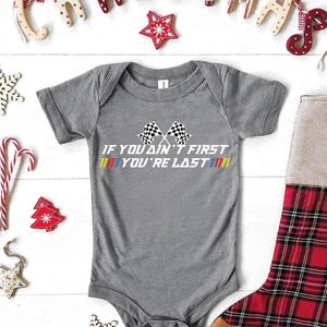 Op de afbeelding: Een grijze babybody met de tekst "IF YOU AIN'T FIRST YOU'RE LAST" en racevlaggen. Kerstversieringen zijn onder meer een rood-zwarte geruite kous, een "CHRISTMAS" banner en ster- en sneeuwvlokornamenten.