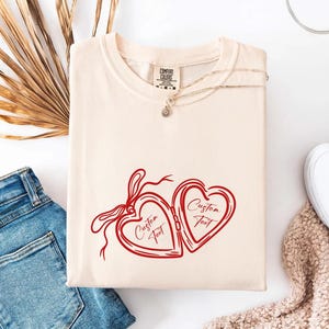 May include: Cream-colored t-shirt with a red heart-shaped locket design. The locket is open, with the words "Custom Text" inside each heart. A delicate necklace rests on the shirt, and blue jeans and white shoes are in the background.