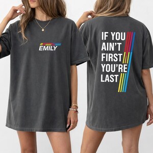 Op de afbeelding: Donkergrijs t-shirt met de naam "EMILY" in witte letters en een kleurrijk streepontwerp. De achterkant van het shirt heeft de tekst "IF YOU AIN'T FIRST YOU'RE LAST" met een verticaal streepontwerp in rood, blauw, geel en groen.