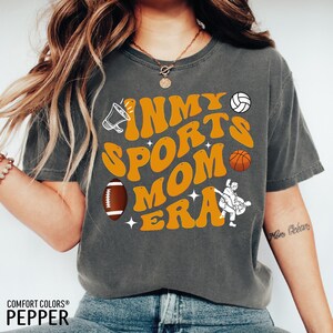 Op de afbeelding: Een grijs T-shirt met de tekst "In My Sports Mom Era" in oranje en witte letters. Het shirt heeft verschillende sportpictogrammen, waaronder een voetbal, een basketbal, een volleybal en een fluitje.