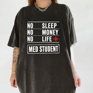 Op de afbeelding: Een antracietgrijs t-shirt met korte mouwen en witte tekst die luidt NO SLEEP, NO MONEY, NO LIFE met een rood plusteken, gevolgd door de tekst MED STUDENT in een wit rechthoekig kader. Het t-shirt heeft een ronde hals en een ruime pasvorm.