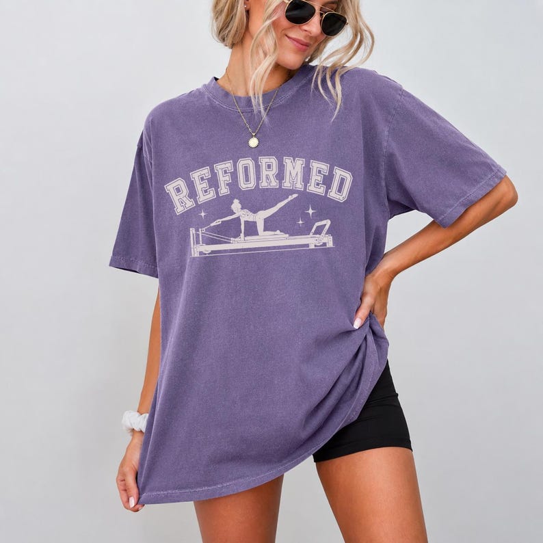 A woman wearing a purple t-shirt with a graphic design and sunglasses posing against a white background.