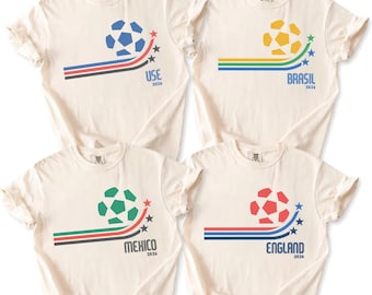 Puede incluir: Cuatro camisetas color crema con gráficos de balón de fútbol y nombres de países. Cada camiseta presenta un balón de fútbol de diferente color, una línea curva y el año 2026. Los países son EE. UU., Brasil, México e Inglaterra.