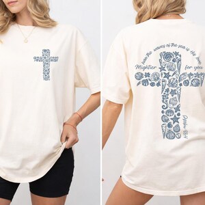 May include: Cream-colored t-shirt with a cross design on the front and back. The cross is filled with blue seashell illustrations. The back of the shirt has the text "han he waves of the sea is the love Mightier for you Psalm 93:4."