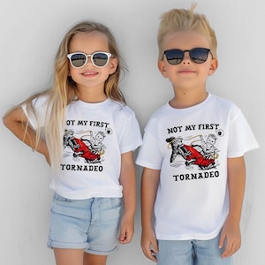 May include: Two children wearing white t-shirts with the text "NOT MY FIRST TORNADEO" and a graphic of a red car in a tornado. The girl wears light blue denim shorts and sunglasses. The boy wears sunglasses.