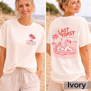 May include: Ivory-colored t-shirt with a pink graphic of palm trees and the text "Emily Bach" on the front. The back features a pink flamingo float with a woman, palm trees, and the text "Last Toast on the Coast".