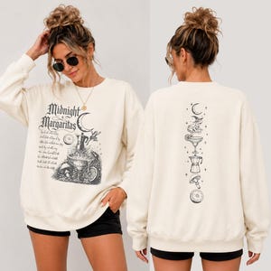 May include: A cream-colored long-sleeve sweatshirt featuring black graphic prints. The front displays the text Midnight Margaritas with a crescent moon, a cocktail glass, a coffee pot, and a frog. The back features a vertical stack of these icons.