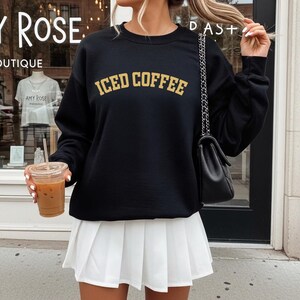 May include: Black sweatshirt with the text "ICED COFFEE" in gold lettering. The sweatshirt is paired with a white pleated skirt. The person is holding a clear plastic cup of iced coffee.