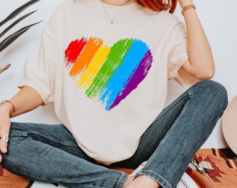 May include: Cream-colored t-shirt featuring a large, colorful heart design. The heart is composed of broad brushstrokes in rainbow colors: red, orange, yellow, green, blue, and purple. The shirt is paired with blue jeans.