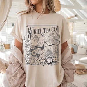 May include: Cream-colored t-shirt featuring a graphic design for Suriel Tea Co. The design includes a teapot, flowers, and the text "The hottest tea in Prythian." The shirt has short sleeves and a relaxed fit.