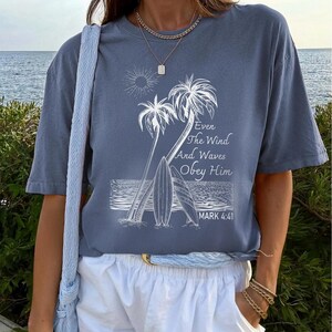 A woman wearing a blue t-shirt with a graphic design of palm trees and waves, standing near a body of water.