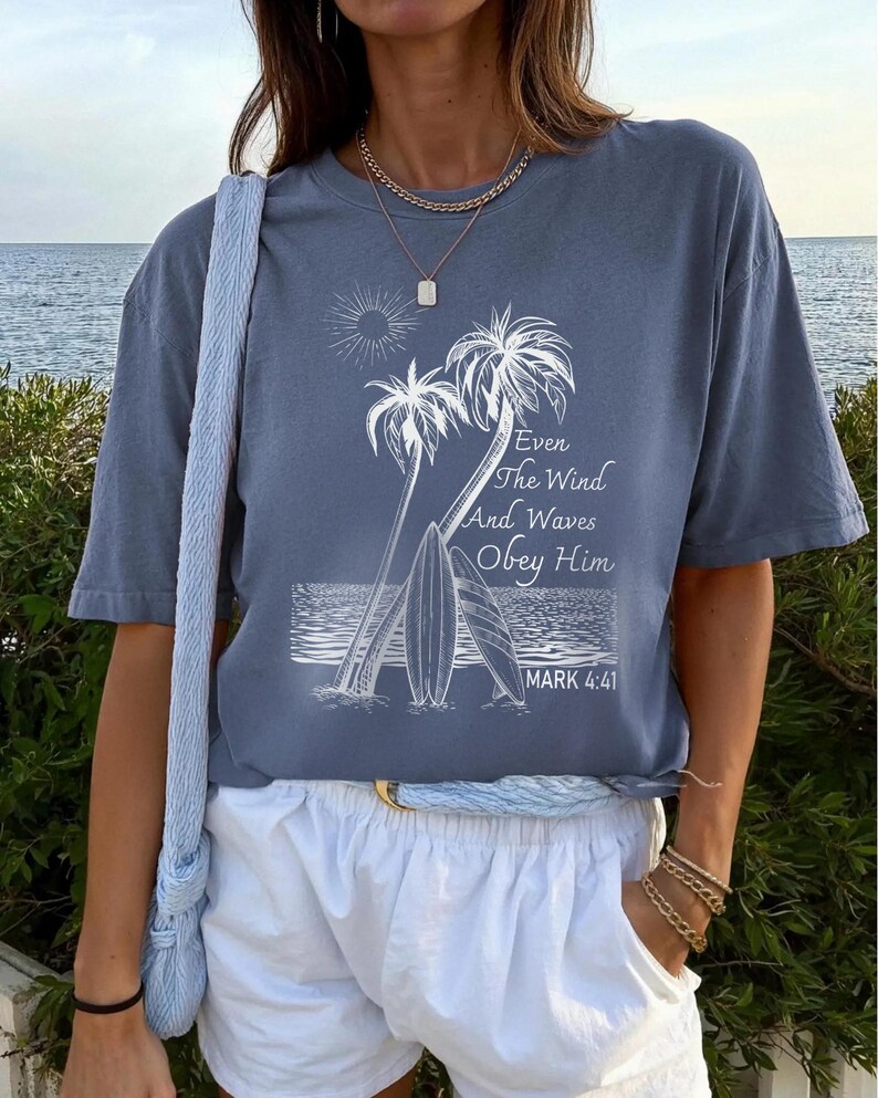 A woman wearing a blue t-shirt with a graphic design of palm trees and waves, standing near a body of water.