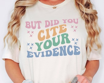 May include: Cream-colored t-shirt with the text "BUT DID YOU CITE YOUR EVIDENCE" in a retro font. The text is in various colors, including pink, orange, green, and blue. The shirt has a relaxed fit and short sleeves.