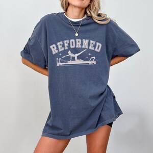 A woman wearing a blue t-shirt with the text "REFORMED" printed on it, along with a graphic of a person doing a Pilates reformer workout.