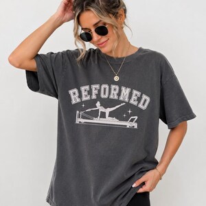 A woman wearing a gray t-shirt with the word "REFORMED" printed on it, posing with her hand on her head.