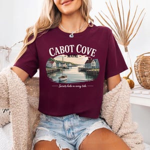 A woman wearing a burgundy t-shirt with a graphic of Cabot Cove, Maine, and the text "Secrets hide in every tide" printed on it.