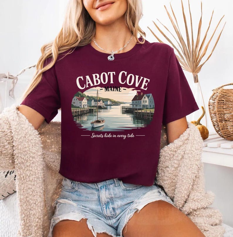 A woman wearing a burgundy t-shirt with a graphic of Cabot Cove, Maine, and the text "Secrets hide in every tide" printed on it.