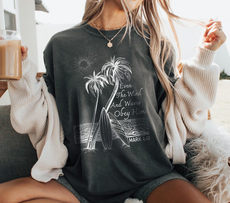 A woman wearing a gray t-shirt with a graphic design of palm trees and a beach scene, holding a drink in her hand.