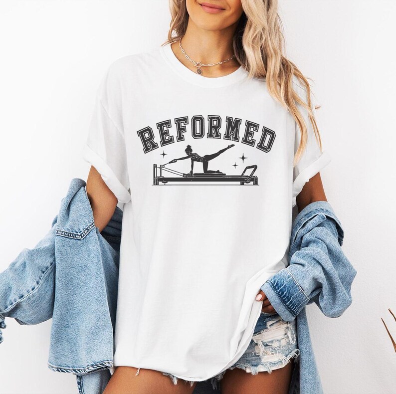 A woman wearing a white t-shirt with the text "REFORMED" and a graphic of a person on a Pilates reformer machine.