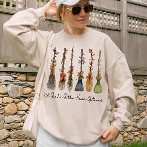 May include: A cream-colored crewneck sweatshirt featuring a graphic of six distinct witch brooms decorated with autumn leaves and orange bows. Below the brooms, the text reads, A Girl's Gotta Have Options, in a black cursive script font.
